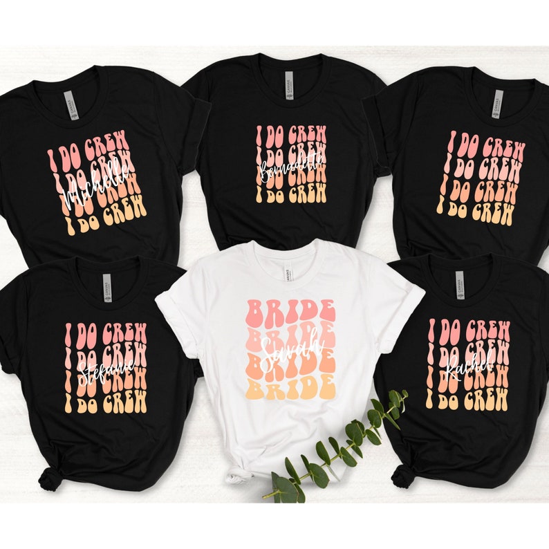 May include: Five t-shirts with a bridal party theme. Four black t-shirts with the text "I Do Crew" in a retro font with a gradient of pink and orange. One white t-shirt with the text "Bride" in a retro font with a gradient of pink and orange.