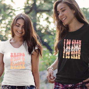 May include: Two women wearing white and black t-shirts with text in a retro font. The white shirt says "Bride Sarah Bride" and the black shirt says "I Do Crew" repeated three times.