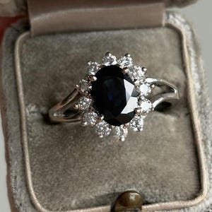 May include: A silver ring with a dark blue oval gemstone, surrounded by smaller white gemstones, displayed in a gray velvet ring box. The ring's band splits into two delicate strands on either side of the central stone.