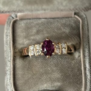 Op de afbeelding: Een gouden ring met een ovale robijn en diamantaccenten, gepresenteerd in een grijze fluwelen ringdoos. De ring heeft een klassiek ontwerp met een gepolijste gouden band en sprankelende edelstenen.