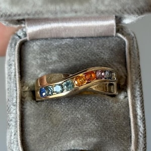 Könnte beinhalten: Ein goldener Ring mit einem Regenbogen aus Edelsteinen, präsentiert in einer grauen Samt-Ringbox. Der Ring hat ein Wellendesign mit einer Reihe farbiger Steine, darunter Blau, Grün, Gelb, Orange und Lila. Die Box ist geöffnet und zeigt die Details des Rings.