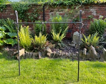 Puede incluir: Un soporte de jardín de metal negro con dos postes verticales y una barra horizontal. El soporte está en un jardín con hierba verde y helechos.