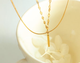 Puede incluir: Un collar de cadena de oro con una cadena fina y delicada y un colgante rectangular simple. El collar se muestra sobre una superficie blanca.