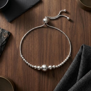 May include: A silver-toned bracelet featuring a series of polished spherical beads of varying sizes, with a larger central bead. The bracelet is constructed with a flexible box chain and includes an adjustable sliding bead closure mechanism.