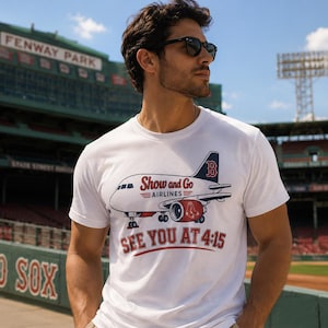 May include: A white short-sleeved t-shirt featuring a graphic of an airplane with the text Show and Go AIRLINES and SEE YOU AT 4:15. The airplane graphic includes a red Boston Red Sox logo on the engine and a blue B on the tail fin.