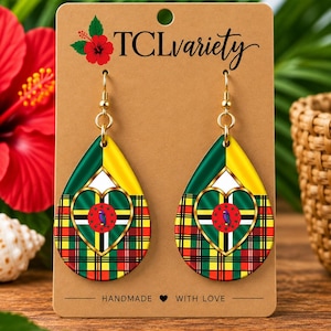 Op de afbeelding: Druppelvormige oorbellen met de vlag van Dominica in een goudomrand hart, geplaatst tegen een groen, geel en rood ruitpatroon. De oorbellen hangen aan goudkleurige haakjes op een bruine displaykaart met de tekst TCLvariety.