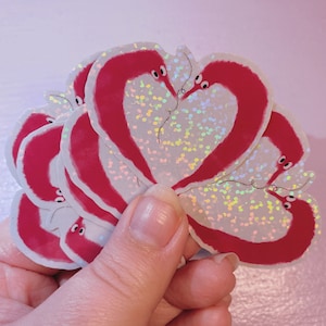 May include: A selection of stickers depicting red worm designs in a heart shape. The stickers have a shimmering, glittery finish and are held in a hand against a pale pink backdrop. The worms have small, black eyes.