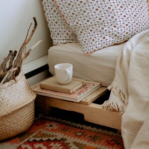 Puede incluir: Una escena de dormitorio acogedora con almohadas estampadas, una manta blanca y una cesta tejida. Una taza blanca descansa sobre una pila de libros en un palé de madera, con una alfombra colorida en primer plano. Las almohadas tienen un fondo blanco con patrones geométricos marrones.