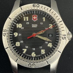 May include: A Swiss Army wristwatch with a black textured dial, silver-tone Arabic numerals, and a rotating bezel with engraved minute markers. The watch features a red second hand, luminous hour and minute hands, a date window, and a black strap.