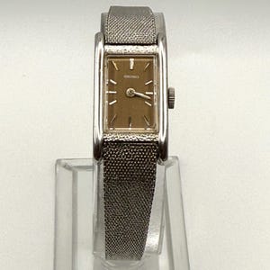 May include: A vintage Seiko wristwatch with a rectangular gold-toned face and silver-toned metal mesh band. The watch has a small crown and a classic, minimalist design. The watch is displayed on a clear stand.