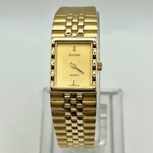 Puede incluir: Un reloj de cuarzo Bulova dorado con una esfera rectangular y un brazalete de eslabones a juego. La esfera del reloj es de color oro claro con el nombre de la marca "Bulova" y la palabra "Quartz" impresa. El reloj tiene un diseño clásico y elegante.