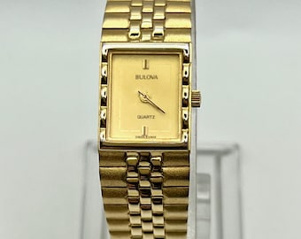 May include: A gold-toned Bulova quartz watch with a rectangular face and a matching link bracelet. The watch face is a light gold color with the brand name "Bulova" and the word "Quartz" printed on it. The watch has a classic, elegant design.