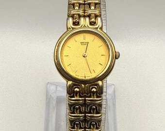 May include: A gold-toned Seiko Quartz watch with a round face and a matching gold link bracelet. The watch face is a light gold color with black hour markers and hands. The watch is displayed on a clear stand.