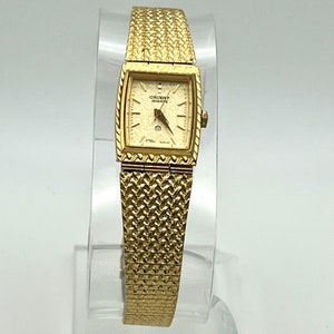 Puede incluir: Un reloj de pulsera Orient Quartz de tono dorado con una esfera cuadrada y una pulsera de metal dorado. La esfera del reloj tiene un fondo blanco con marcas negras y las palabras "Orient Quartz" impresas en ella.