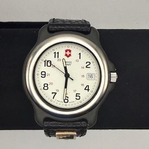 May include: A Swiss Army wristwatch featuring a round silver-tone metal case, a white dial with black Arabic numerals, a date window at the three o'clock position, and a black textured leather strap with a small metallic accent near the buckle.