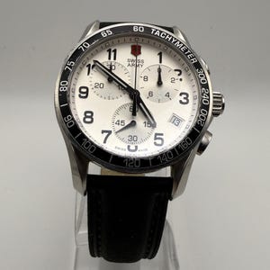 May include: A Swiss Army chronograph watch with a white dial, black Arabic numerals, and three sub-dials. The watch features a black tachymeter bezel, a date window at the four o'clock position, black hands, and a black leather strap with stitching.