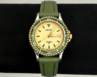 May include: A Seiko Sports 50 wristwatch with a gold-toned face, a green bezel, and a green leather band. The watch has a date display at the 3 o'clock position and gold-toned hands. The watch face has the Seiko logo and the words "Sports 50" printed on it.
