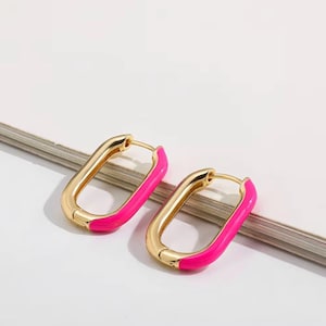 Può includere: Un paio di orecchini a cerchio color oro di forma rettangolare. L'interno dei cerchi è dipinto con smalto rosa acceso. Gli orecchini sono esposti su una superficie bianca.