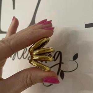 Può includere: Un anello color oro caratterizzato da un design a spirale multistrato con quattro fasce distinte, lisce e arrotondate impilate insieme. Il metallo presenta una finitura lucida e altamente riflettente che cattura la luce in modo uniforme.