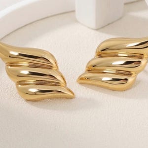 Può includere: Un paio di orecchini color oro con finitura lucida e liscia, caratterizzati da un design a strati simile ad ali. Ogni orecchino è composto da tre segmenti distinti, curvi e arrotondati che si restringono verso un'estremità.