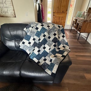 May include: A handmade quilt with a geometric pattern of squares in shades of blue, black, and white is draped over a black leather sofa. The quilt's design creates a visually appealing texture, adding a touch of comfort and style to the living room.