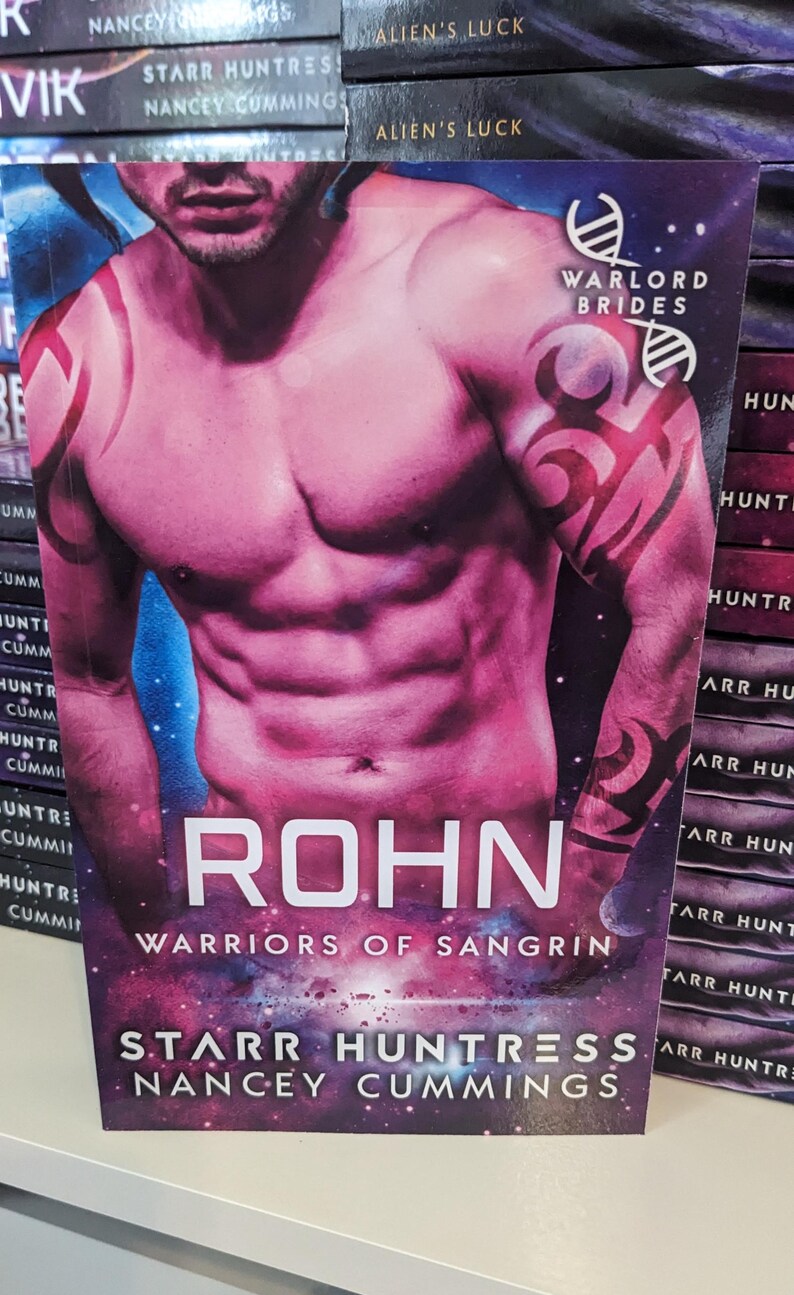 May include: A paperback book cover with a muscular man on the front. The cover is pink and purple with a galaxy background. The title of the book is "ROHN: WARRIORS OF SANGRIN" by Starr Huntress and Nancy Cummings.