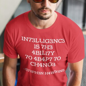 May include: Red t-shirt with white text that reads "INT3LLIG3NC3 15 7H3 4B1L1TY 70 4B4P7 70 CH4NG3 -573PH3N H4WK1NS"