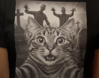 May include: Black t-shirt featuring a graphic of a cat taking a selfie with zombies in a graveyard. The cat has wide eyes and an open mouth. The background is dark with lit candles.