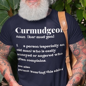 May include: Navy blue t-shirt with the definition of "Curmudgeon" printed in white. The definition includes the word's pronunciation and a description of a person who is easily annoyed. The shirt is a crew neck.