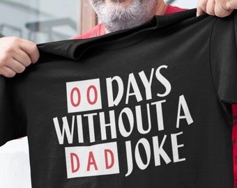 Könnte beinhalten: Schwarzes T-Shirt mit dem weißen Text "00 DAYS WITHOUT A DAD JOKE", wobei die Zahlen und das Wort "DAD" in roten Kästchen stehen. Das T-Shirt wird von einer Person hochgehalten.