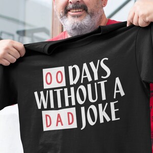 May include: Black t-shirt with the text "00 DAYS WITHOUT A DAD JOKE" in white, with the numbers and the word "DAD" in red boxes. The shirt is being held up by a person.