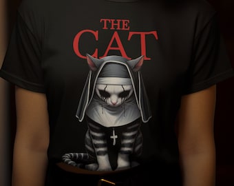 May include: A black short-sleeved t-shirt featuring a graphic print of a cat dressed in a nun's habit with a cross necklace. The text above the cat reads THE CAT in red capital letters. The cat has dark markings around its eyes and ears.