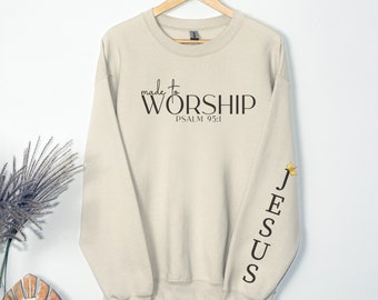 Puede incluir: Una sudadera beige con el texto "made to WORSHIP PSALM 95:1" en la parte delantera y "JESUS" con una estrella dorada en la manga.