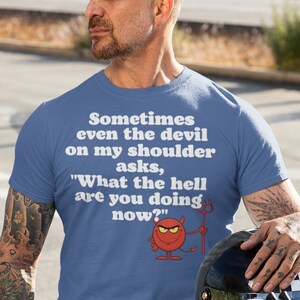 May include: A man wearing a blue t-shirt with a white graphic that says "Sometimes even the devil on my shoulder asks, "What the hell are you doing now?" with a red cartoon devil holding a pitchfork. He is sitting on a black motorcycle.