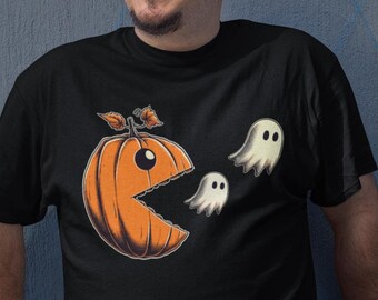 May include: A black short-sleeved t-shirt featuring a graphic print of an orange pumpkin shaped like a video game character with a wide-open mouth, chasing two small white ghosts. The pumpkin has a stem with leaves on top and a single large eye.