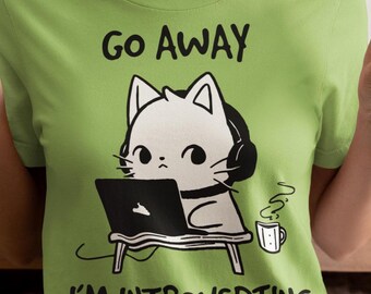May include: Green t-shirt featuring a white cat wearing headphones and using a laptop. The text on the shirt reads "Go Away I'm Introverting".