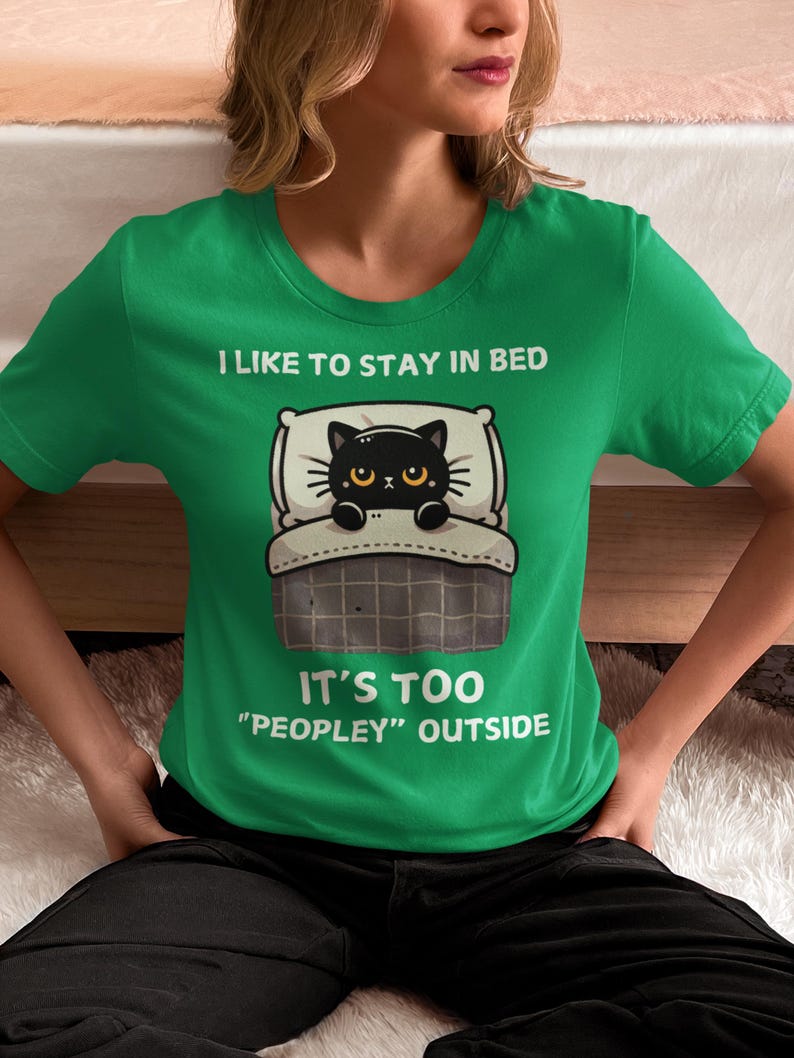 May include: A green t-shirt with a black cat in bed and the text "I like to stay in bed. It's too "peopley" outside."