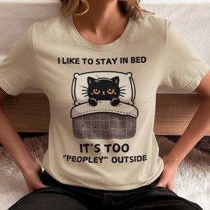 May include: A beige t-shirt with a black cat illustration in bed. The text on the shirt reads "I like to stay in bed. It's too "peopley" outside."