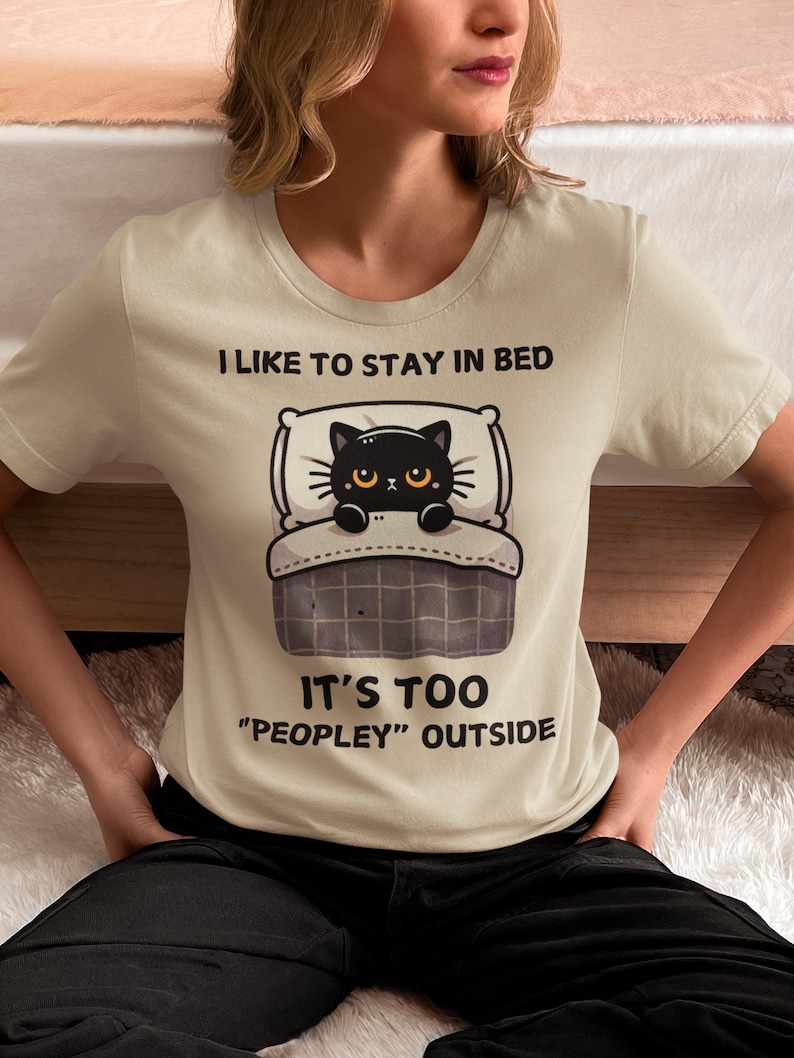 May include: A beige t-shirt with a black cat illustration in bed. The text on the shirt reads "I like to stay in bed. It's too "peopley" outside."