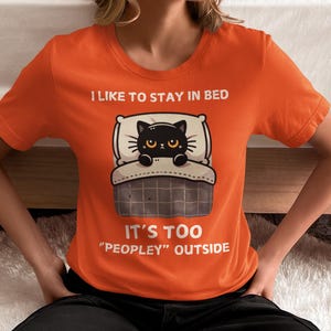 May include: Orange t-shirt with a black cat in bed illustration. The text on the shirt reads "I like to stay in bed. It's too "peopley" outside."