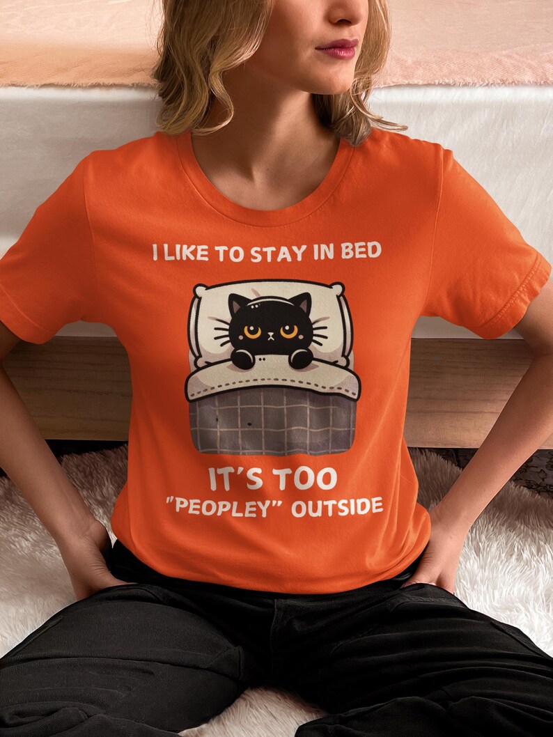 May include: Orange t-shirt with a black cat in bed illustration. The text on the shirt reads "I like to stay in bed. It's too "peopley" outside."