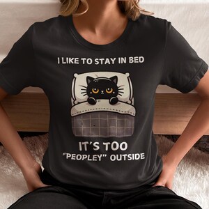 May include: Black t-shirt with a cartoon black cat in bed. The text reads "I like to stay in bed. It's too "peopley" outside."