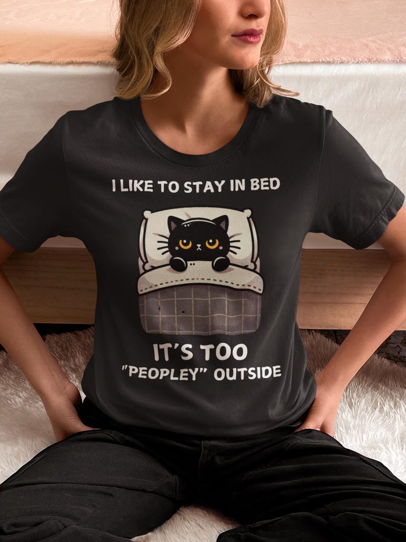 May include: Black t-shirt with a cartoon black cat in bed. The text reads "I like to stay in bed. It's too "peopley" outside."