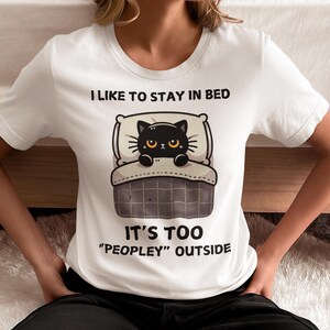 May include: White t-shirt with a black cat in bed graphic. The text on the shirt reads "I like to stay in bed. It's too "peopley" outside."