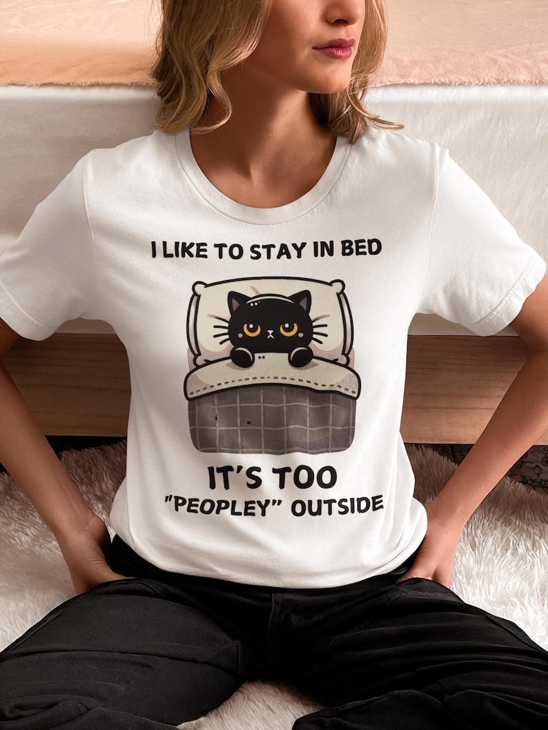 May include: White t-shirt with a black cat in bed graphic. The text on the shirt reads "I like to stay in bed. It's too "peopley" outside."
