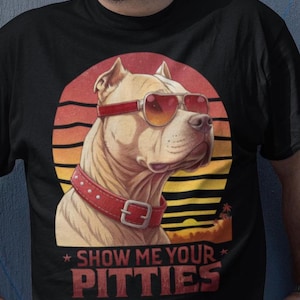 Pode incluir: Camiseta preta com um pitbull usando óculos de sol e coleira vermelha, contra um pôr do sol. O texto "SHOW ME YOUR PITTIES" está impresso em vermelho.