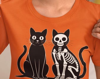 May include: An orange short-sleeved t-shirt featuring a graphic print of two black cats sitting side by side. One cat is solid black, while the other cat is depicted as a white skeleton with a skull face. The design is centred on the chest area.