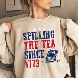 May include: A beige sweatshirt with the text "Spilling the Tea Since 1773" in red, white, and blue. A blue and white ship with three stars is above the text.