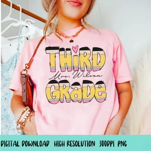 Op de afbeelding: Roze T-shirt met een grafisch ontwerp van het woord "THIRD" in geel en zwart met een roze hart erboven. De tekst "Mrs. Wilson" staat onder het woord "THIRD" en het woord "GRADE" staat eronder. Het grafische ontwerp is gemaakt om eruit te zien als potloden.