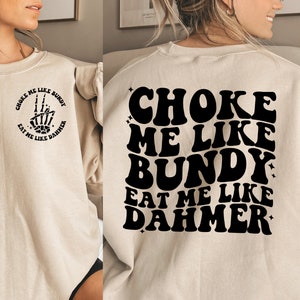 Op de afbeelding: Een beige sweatshirt met zwarte tekst die "CHOKE ME LIKE BUNDY EAT ME LIKE DAHMER" luidt. De tekst is in een retro-lettertype stijl.